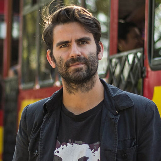 A man with short dark hair and a beard stands outdoors in front of a red and yellow bus, looking at the camera with a neutral expression.