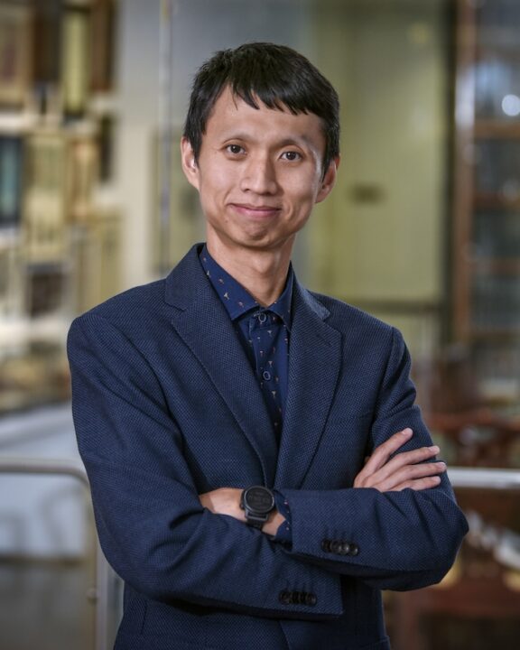 A person wearing a blue suit and patterned shirt stands indoors with arms crossed, smiling at the camera. The background is blurred, showing shelves and framed items.