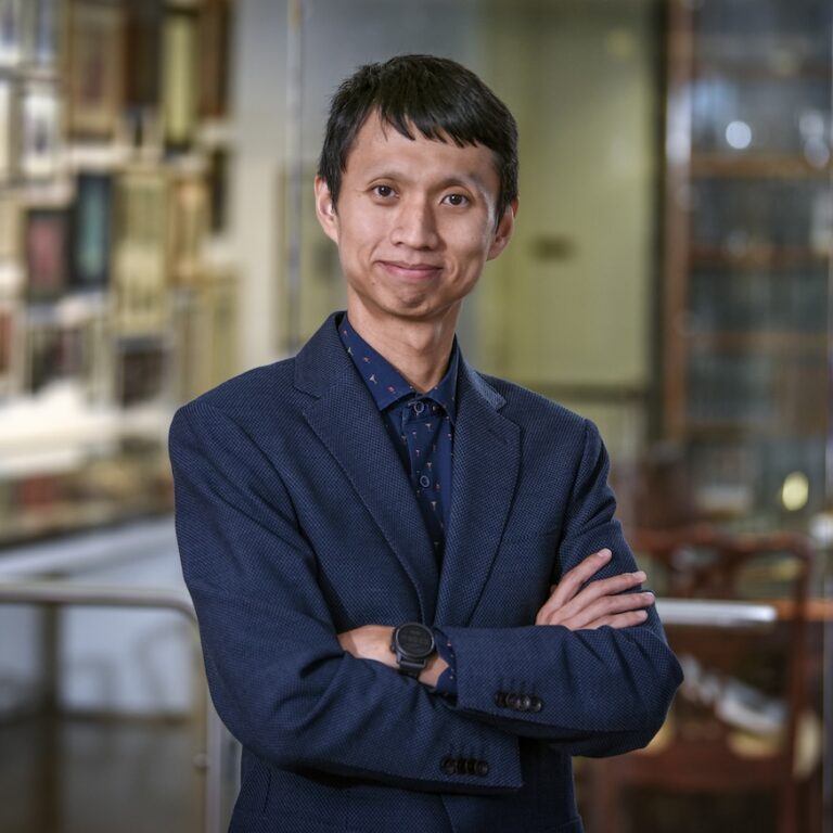 A person wearing a blue suit and patterned shirt stands indoors with arms crossed, smiling at the camera. The background is blurred, showing shelves and framed items.