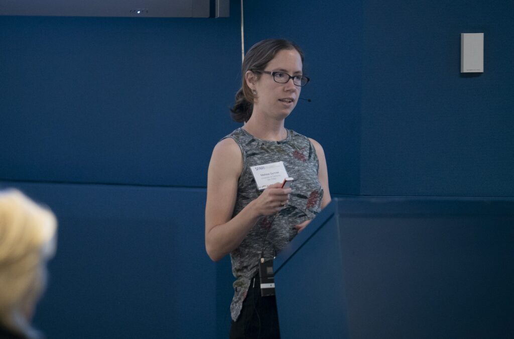 A woman with glasses and a floral sleeveless top speaks at a podium in a blue-walled room, holding a pen and paper with a name tag visible on her chest. A blurred person sits in the foreground.