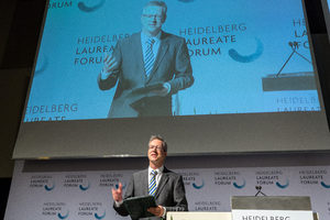 A man in a suit speaks on stage at the Heidelberg Laureate Forum, holding a folder. His large image is projected on a screen above him, both with the events branding in the background.