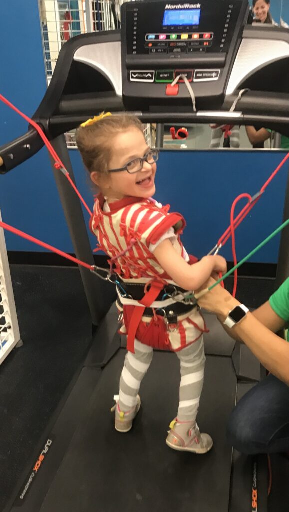 A young girl in a harness stands on a treadmill, smiling at the camera. Colorful cords are attached to her harness, and an adults hand is helping adjust her gear.