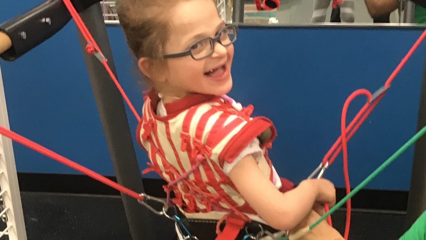 A smiling young girl with glasses is supported by colorful harnesses while standing on a treadmill, with someone adjusting the equipment beside her. The background features a bright blue wall and a mirror.