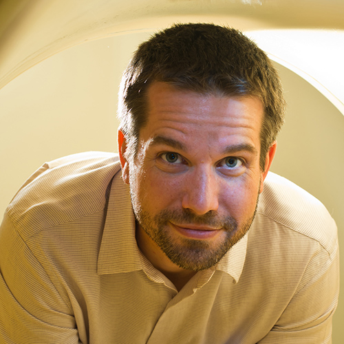 A man with short brown hair, beard, and mustache smiles gently while looking up. He is wearing a beige collared shirt and is illuminated by soft yellowish light.