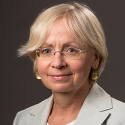 A woman with short blonde hair and glasses wearing a light-colored blazer and gold earrings smiles gently against a plain dark background.