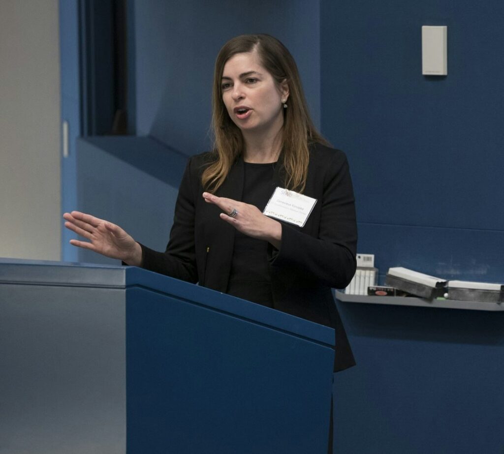 A woman with long brown hair speaks at a podium in a blue room, gesturing with her hands. She wears a black blazer and a name badge.