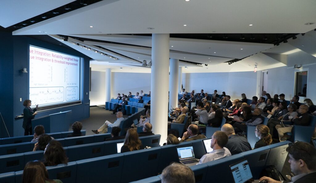 A woman presents slides to an audience in a modern lecture hall; attendees are seated in blue chairs, some taking notes on laptops, while a large screen displays a presentation with text and graphs.