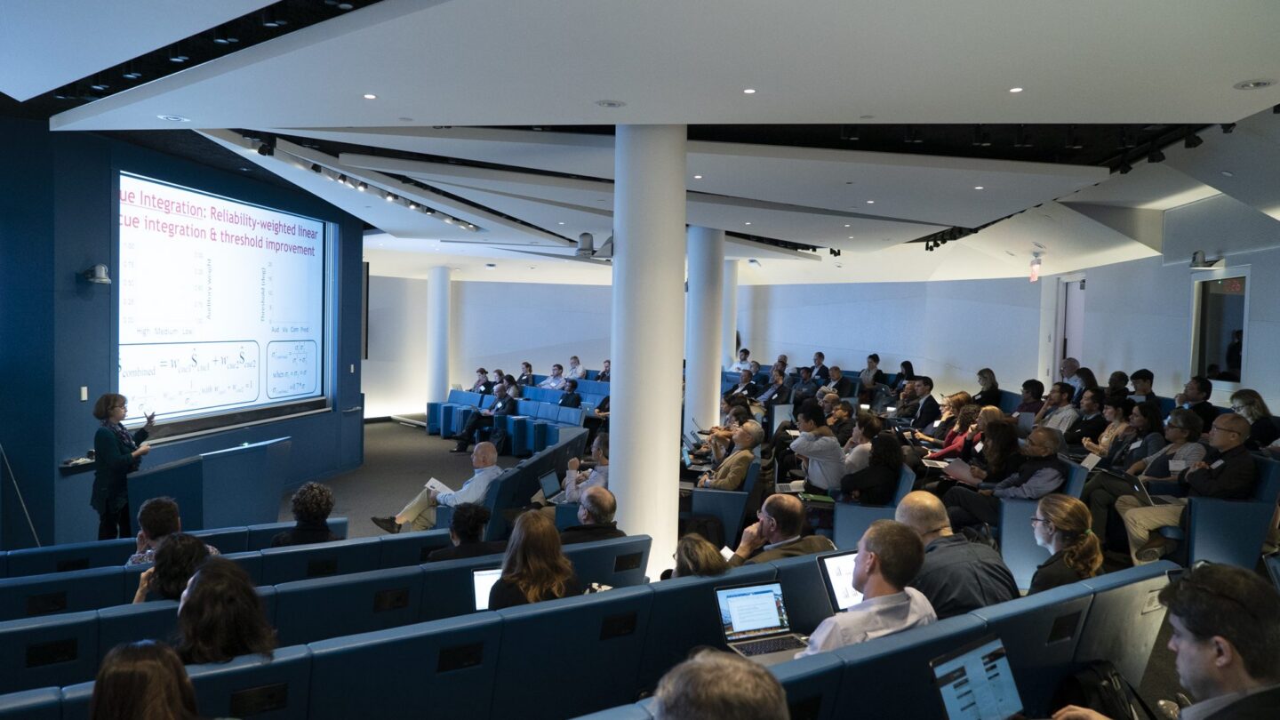 A woman presents slides to an audience in a modern lecture hall; attendees are seated in blue chairs, some taking notes on laptops, while a large screen displays a presentation with text and graphs.