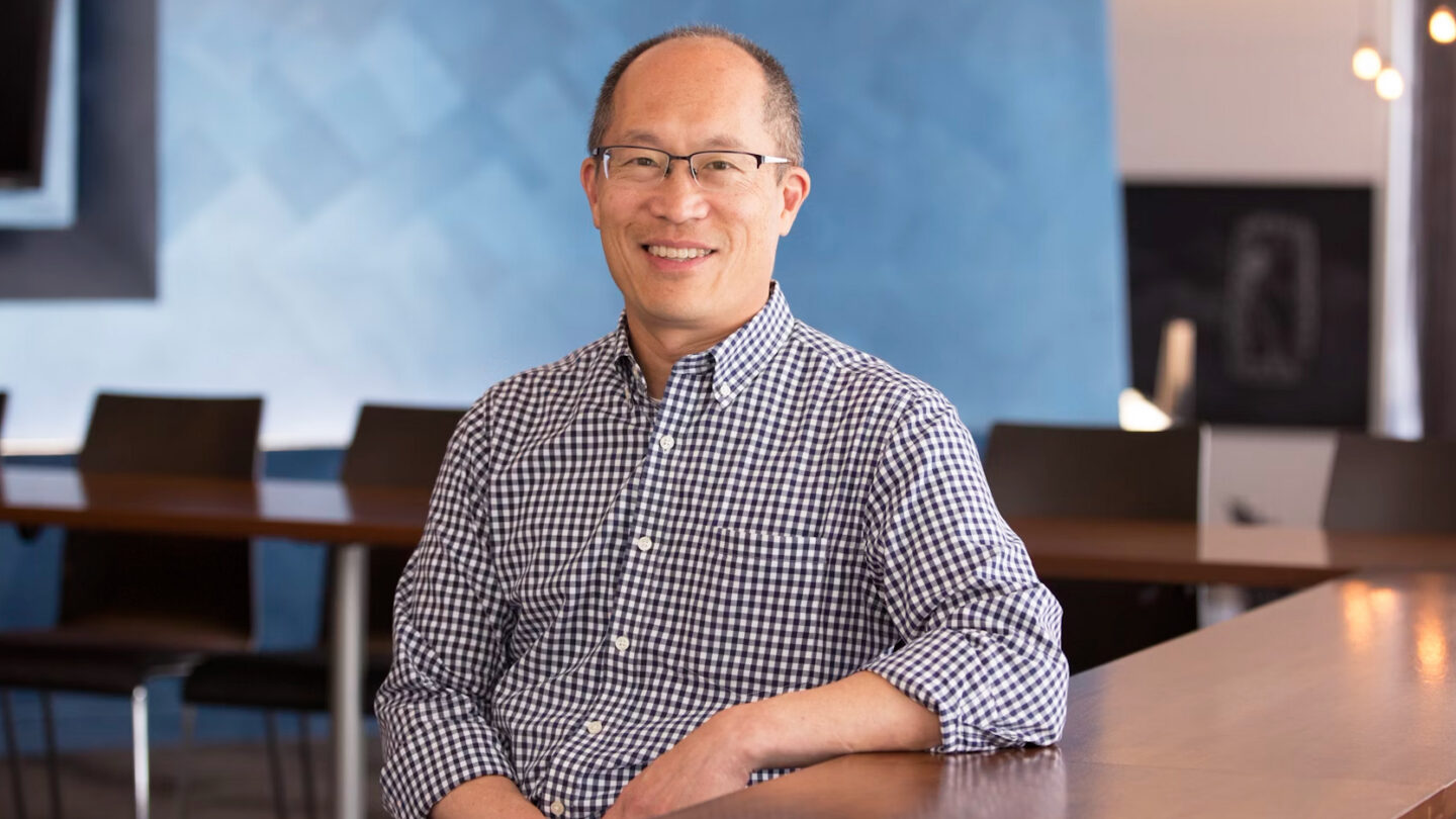 A man wearing glasses and a checkered shirt sits at a table in a modern, well-lit office space with blue walls and empty chairs in the background, smiling at the camera.