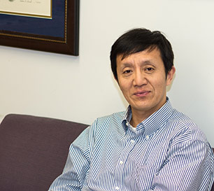 A man with short black hair, wearing a blue and white striped shirt, sits on a purple couch, smiling slightly. A framed document hangs on the light-colored wall behind him.