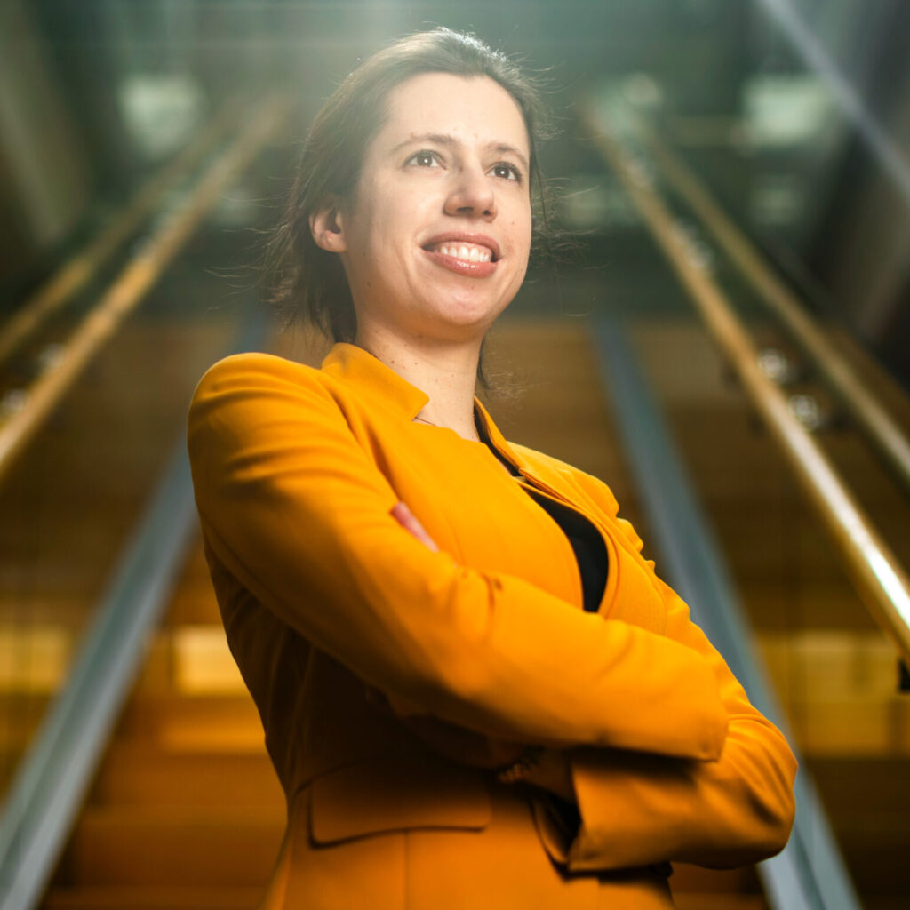 Confident person in a yellow blazer stands with arms crossed in front of an escalator, smiling and looking slightly upward. Bright lighting and blurred background suggest a modern indoor setting.