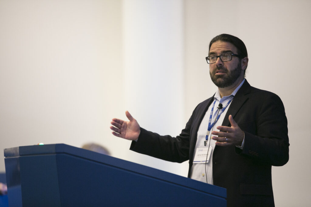 A man with glasses and a beard speaks at a podium, gesturing with both hands. He wears a dark suit jacket and a conference badge, with a light background behind him.