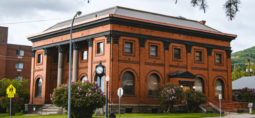 A historic red-brick courthouse with black trim, tall columns, arched windows, and a clock above the entrance, surrounded by green grass, flowering bushes, and street signs.