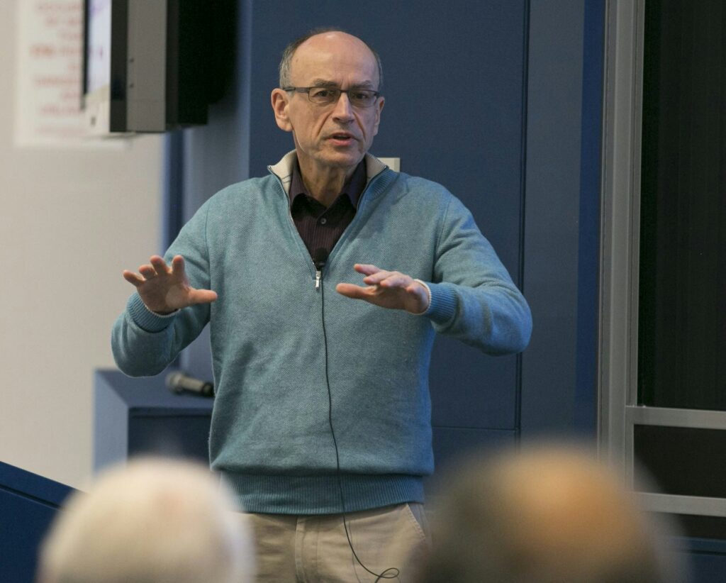 A man wearing glasses and a blue sweater speaks while gesturing with both hands in front of an audience in a lecture room.