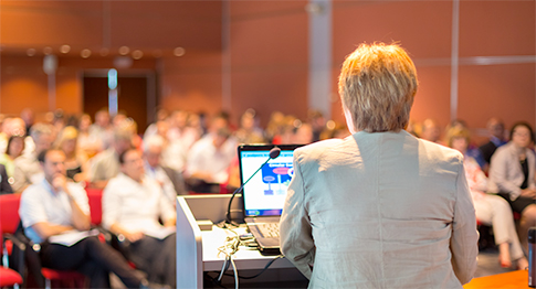 A person speaks at a podium in front of an audience in a conference room, with a laptop displaying a presentation.