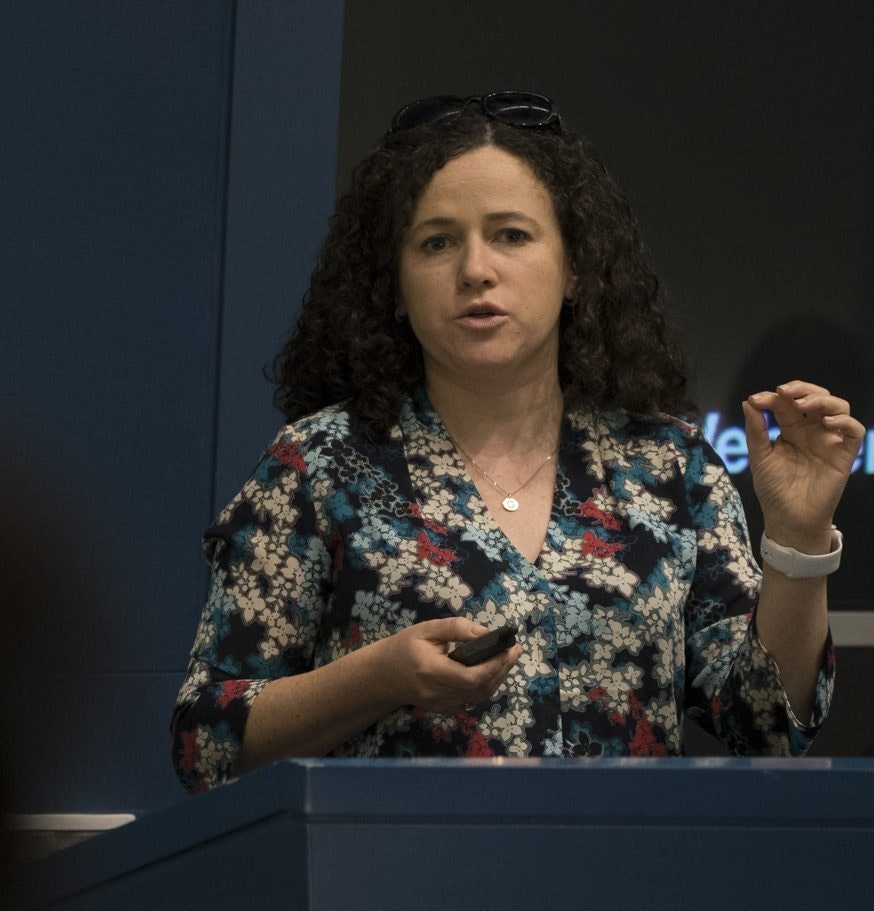 A woman with curly hair, wearing a floral blouse and white watch, speaks while standing behind a podium. She gestures with one hand and holds a remote in the other.