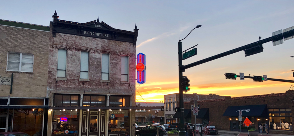 A historic brick building with an R.C. Scripture 1882 sign and a neon Rex sign at sunset, located at a downtown street corner with traffic lights and storefronts.