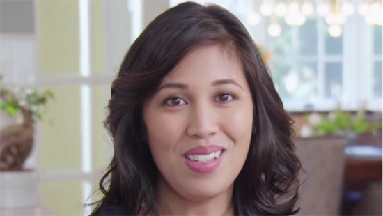 A woman with wavy dark hair smiles at the camera in a bright indoor setting with blurred lights and windows in the background.