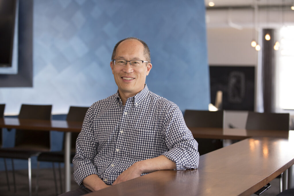 A man wearing glasses and a checkered shirt sits and smiles in a modern office with blue walls and wooden tables.