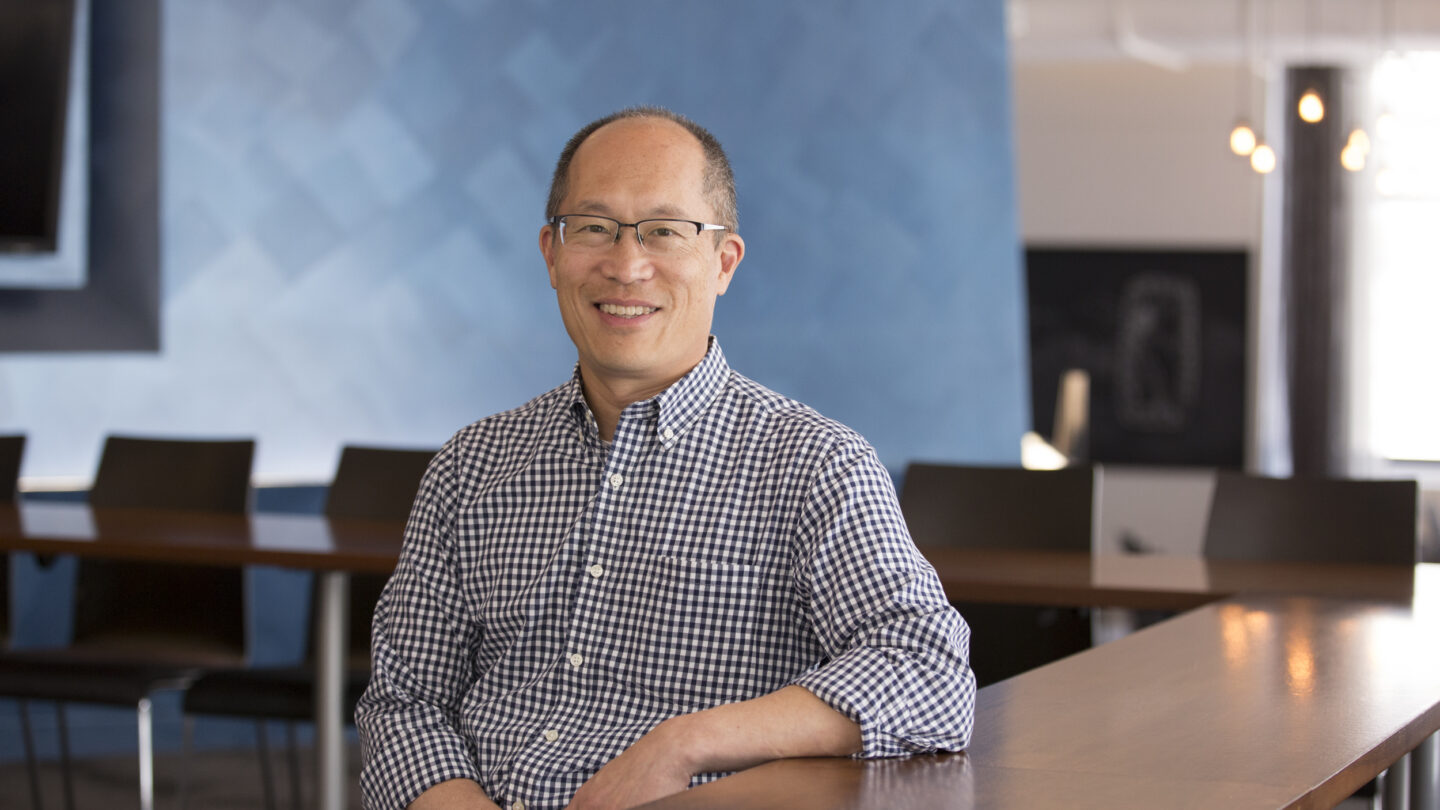 A man wearing glasses and a checkered shirt sits and smiles in a modern office with blue walls and wooden tables.