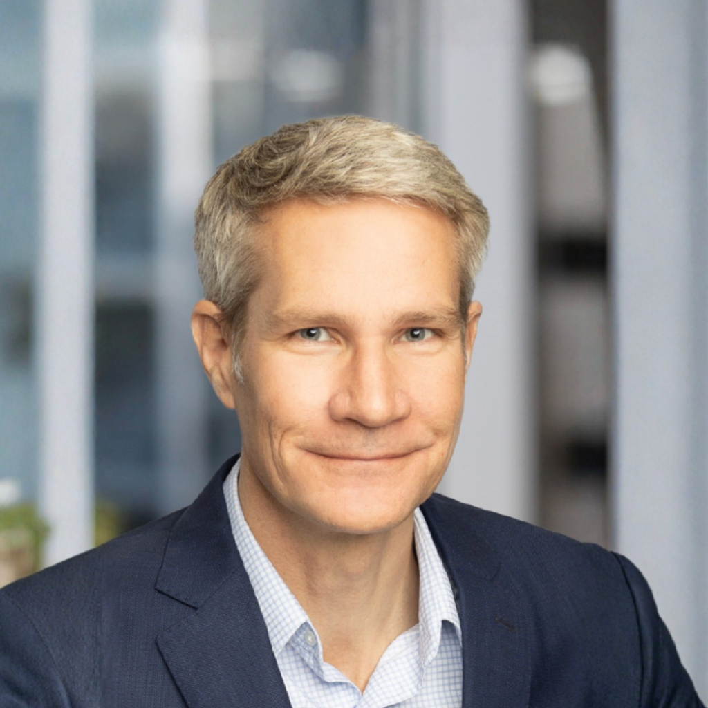 A man with short gray hair wearing a navy blue suit and light blue shirt smiles in an office setting with large windows in the background.