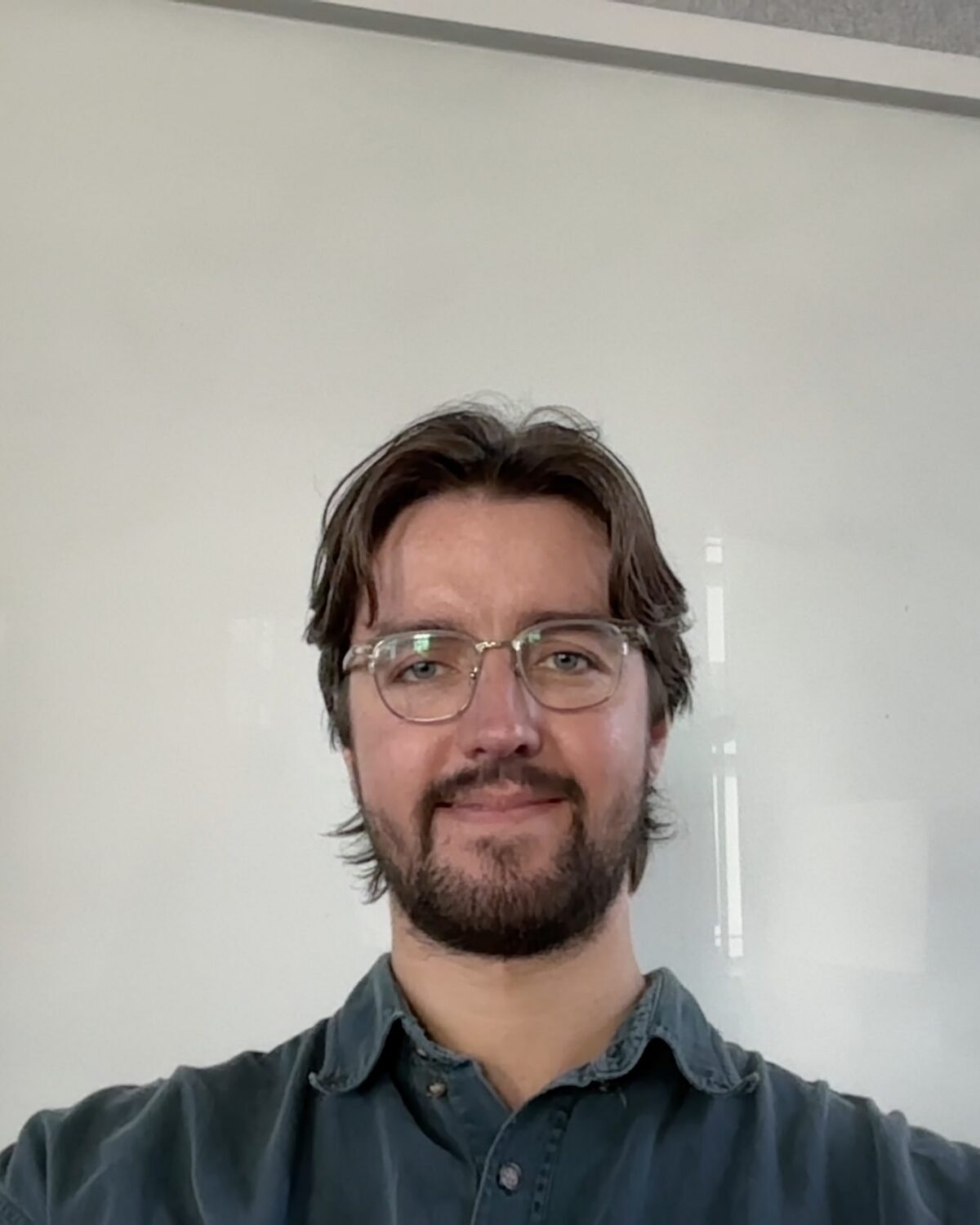 A person with glasses, beard, and medium-length brown hair sits in front of a blank whiteboard, smiling slightly.