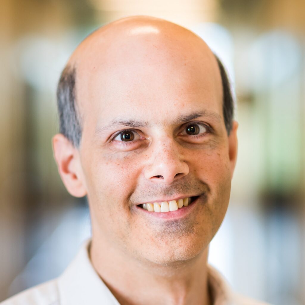 A smiling middle-aged man with a bald head and light skin, wearing a white shirt, is photographed indoors with a softly blurred background.