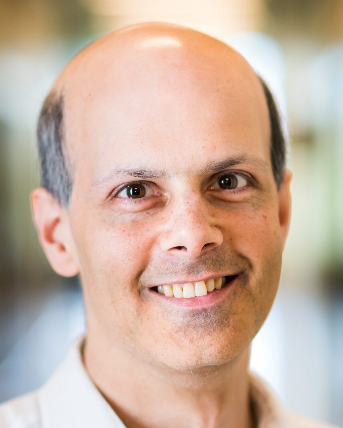 A smiling middle-aged man with a bald head and light skin, wearing a white shirt, is photographed indoors with a softly blurred background.