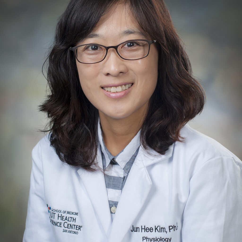 A woman with wavy brown hair, glasses, and a white lab coat smiles at the camera. Her coat reads Jun Hee Kim, PhD, Physiology and has a university logo. The background is softly blurred.