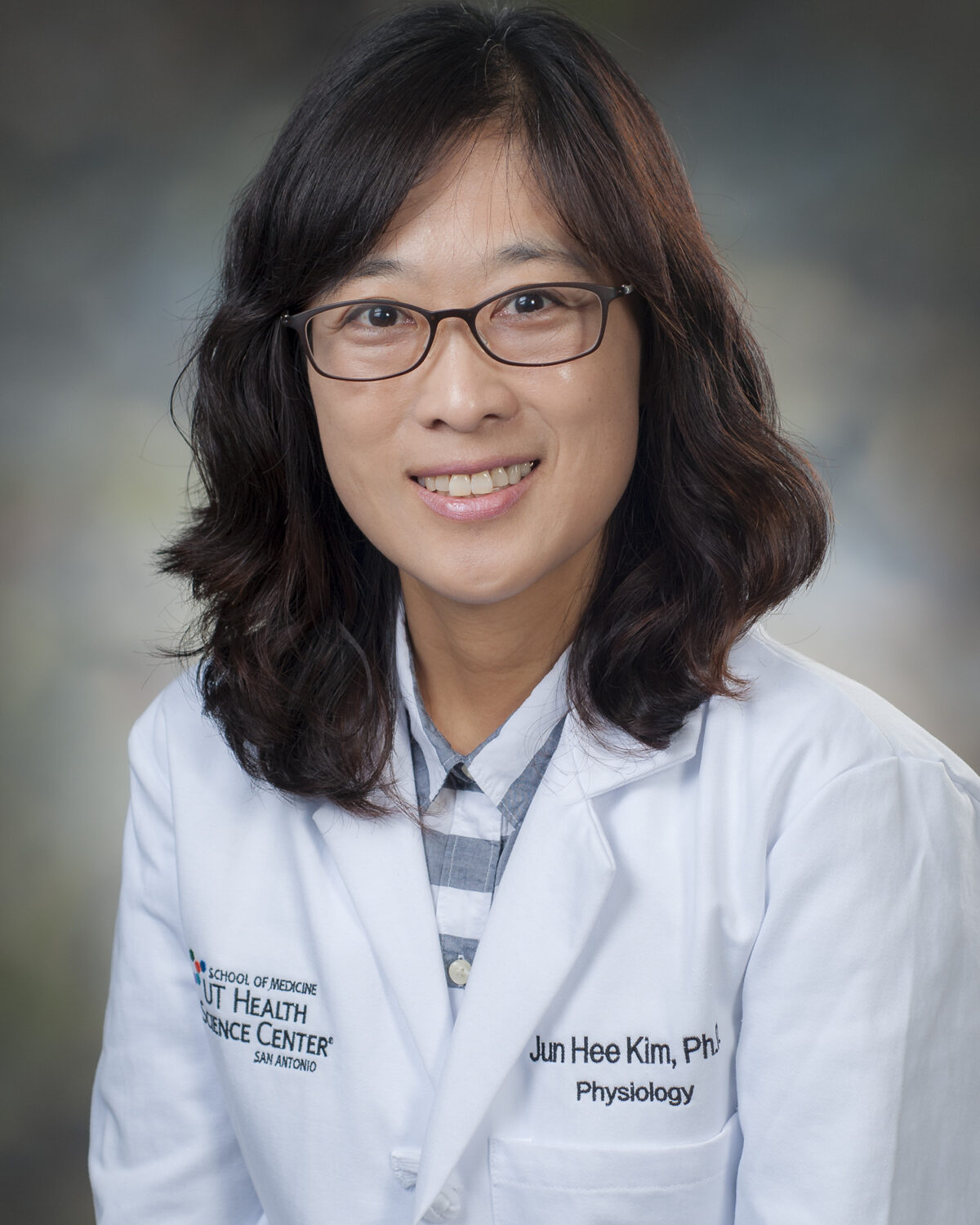 A woman with wavy brown hair, glasses, and a white lab coat smiles at the camera. Her coat reads Jun Hee Kim, PhD, Physiology and has a university logo. The background is softly blurred.