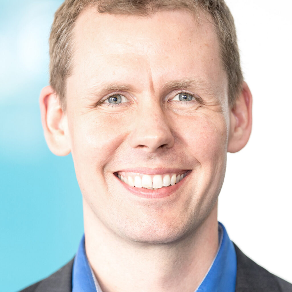 A smiling man with short light brown hair, wearing a blue shirt and a dark blazer, stands in front of a blurred light blue and white background.