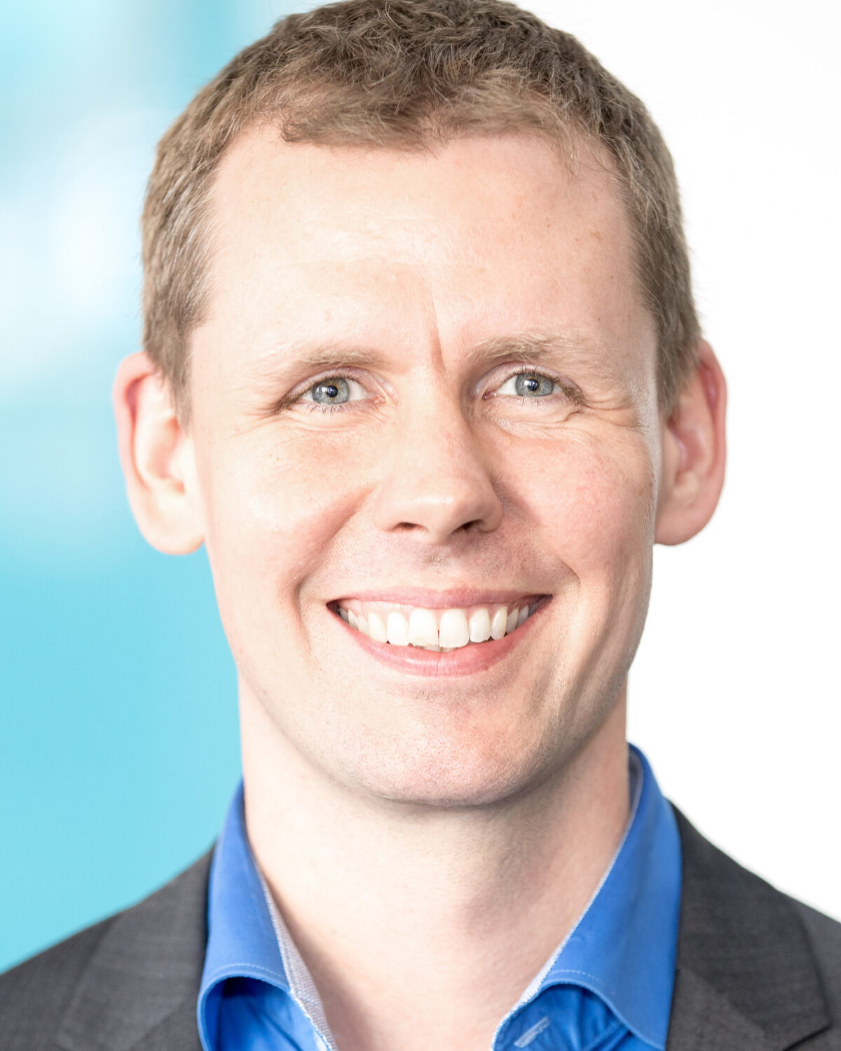 A smiling man with short light brown hair, wearing a blue shirt and a dark blazer, stands in front of a blurred light blue and white background.