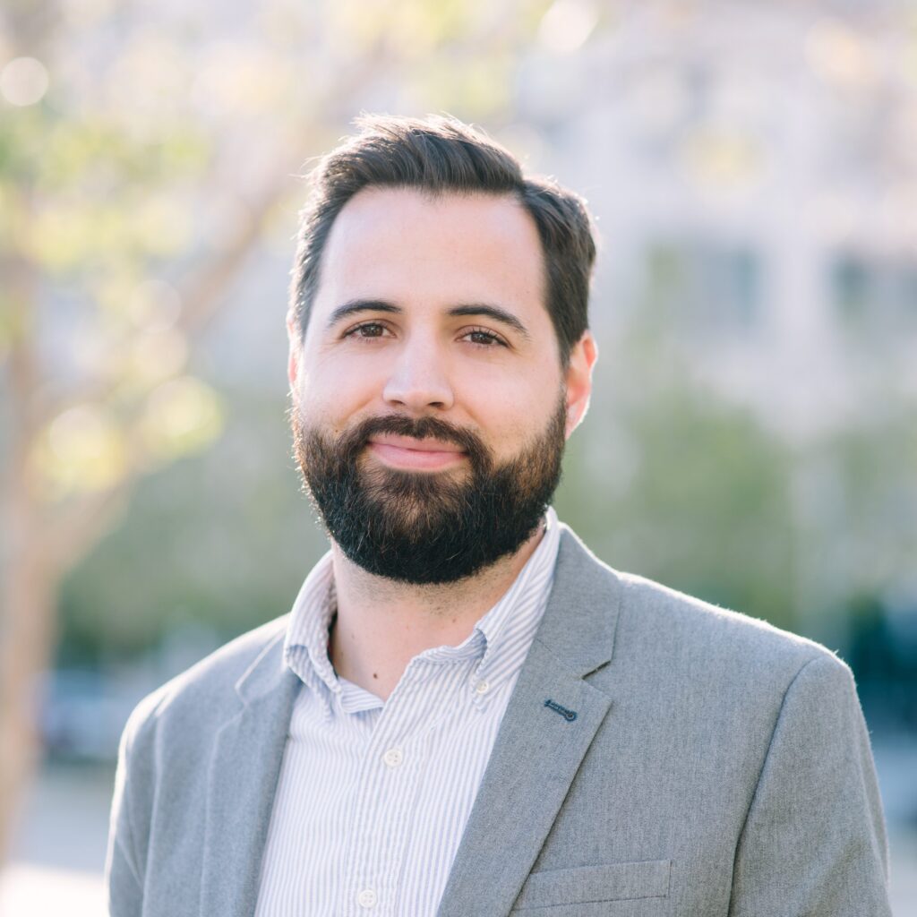 A man with dark hair and a beard, wearing a light gray blazer and striped shirt, stands outdoors, smiling softly with a blurred background of trees and buildings.