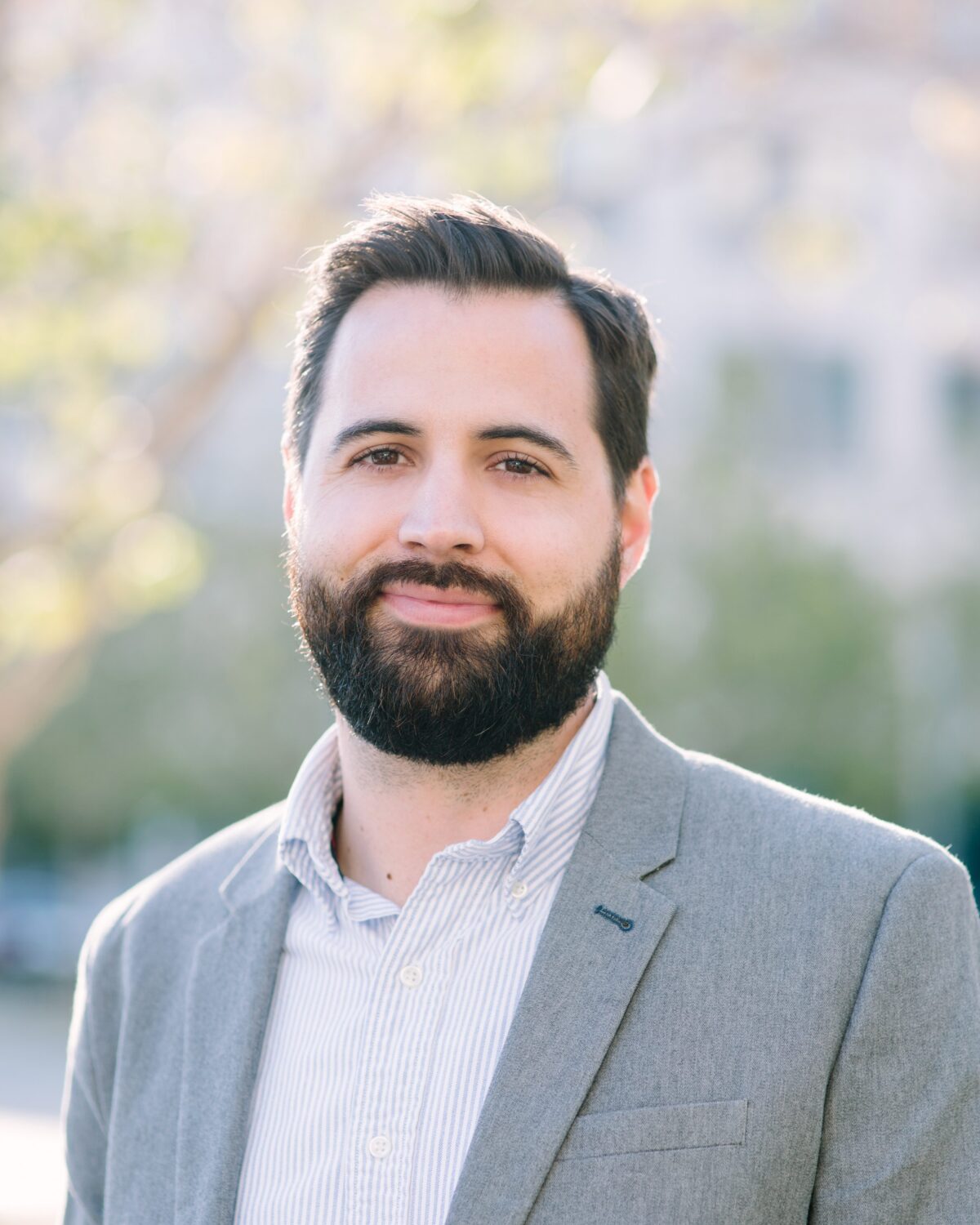A man with dark hair and a beard, wearing a light gray blazer and striped shirt, stands outdoors, smiling softly with a blurred background of trees and buildings.