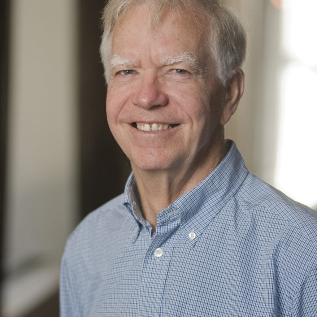 An older man with short white hair, wearing a light blue checked button-up shirt, smiles while standing indoors in soft natural light.