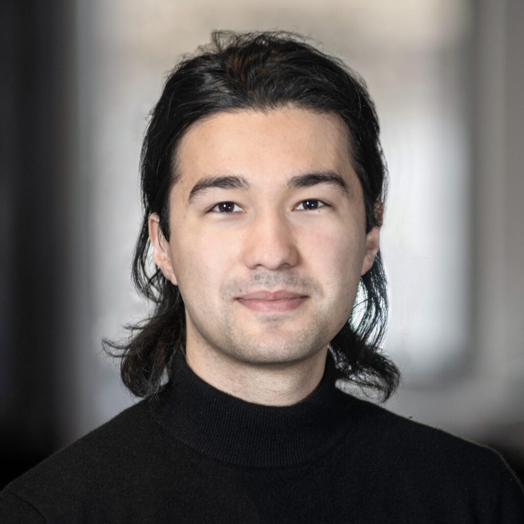 A person with long dark hair, wearing a black turtleneck, smiles softly and looks at the camera. The background is blurred and neutral in color.