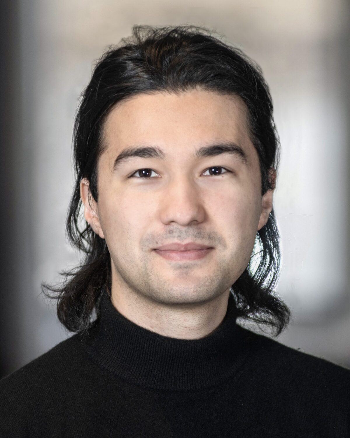 A person with long dark hair, wearing a black turtleneck, smiles softly and looks at the camera. The background is blurred and neutral in color.