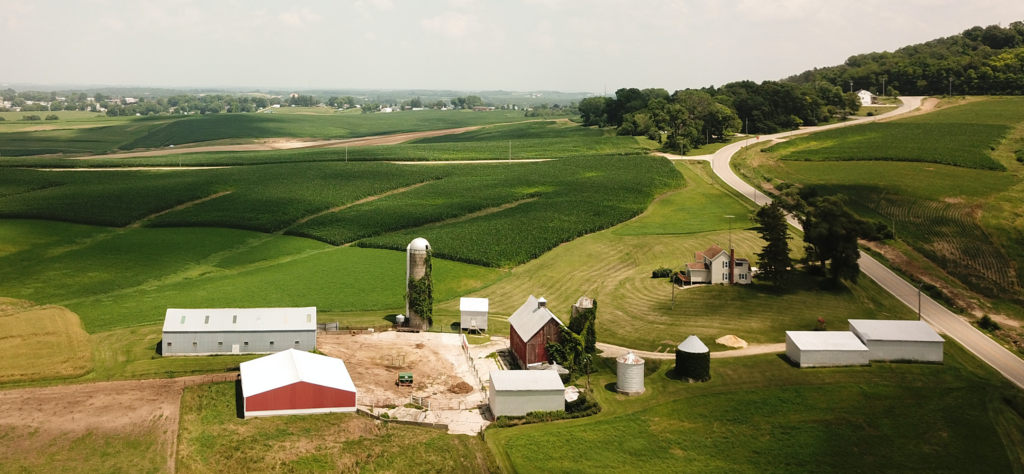 An aerial view of a farmstead with multiple barns, silos, and a house, surrounded by rolling green farm fields and a country road.