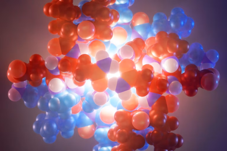 A 3D molecular model with red, blue, and purple spheres clustered together, glowing brightly at the center against a dark background, representing a complex chemical or biological structure.