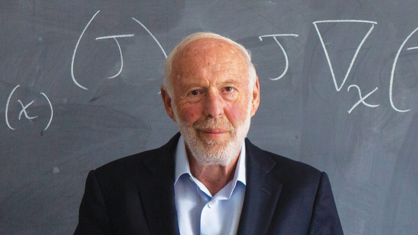 Jim Simons stands in front of a chalkboard. A mathematical equation is written on the board.