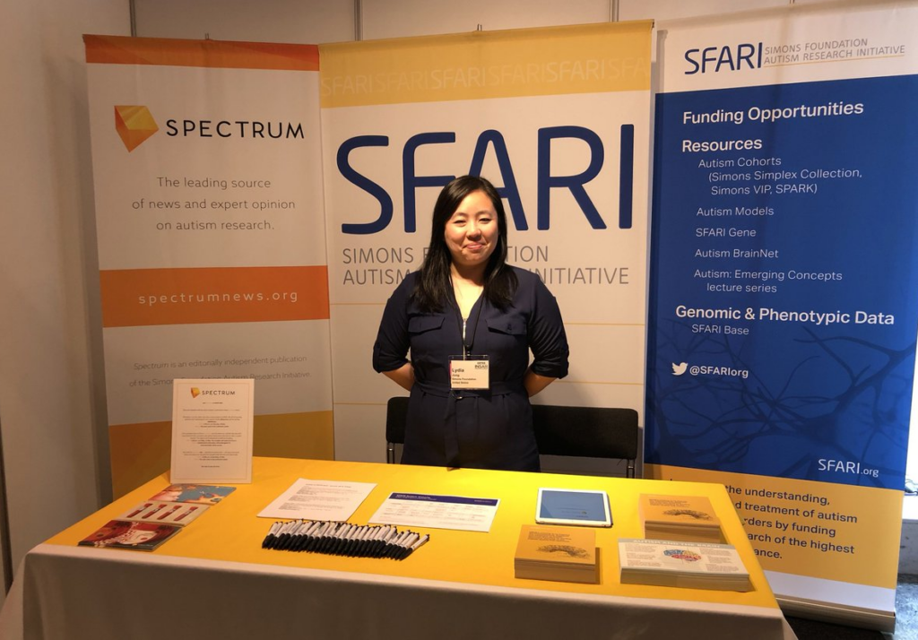 A woman stands at a booth with SFARI and Spectrum banners behind her. The table in front has flyers, pens, and pamphlets about autism research and funding opportunities.