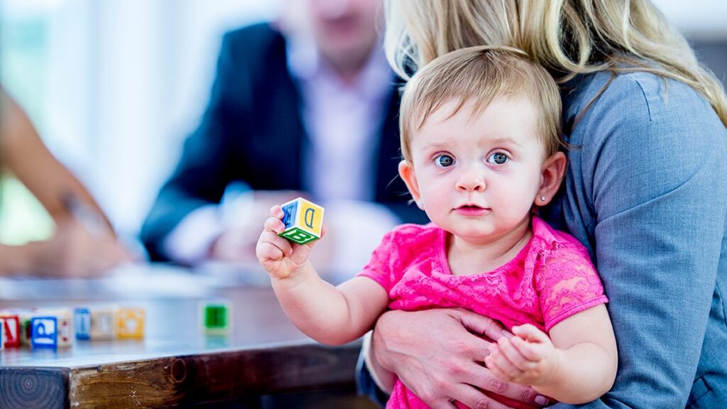 A baby in a pink shirt sits on a womans lap, holding a letter block. The woman hugs the child, and blurry adults are visible in the background at a table.