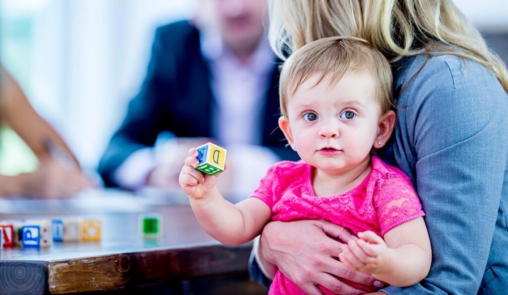 A baby in a pink shirt sits on a womans lap, holding a letter block. The woman hugs the child, and blurry adults are visible in the background at a table.