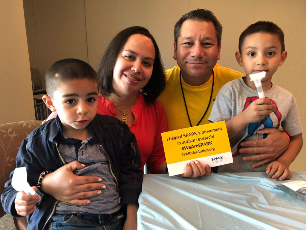 A family of four sits at a table, smiling. The father holds a yellow sign about autism research. The two young boys each hold a swab, and the mother has her arm around one of the children.