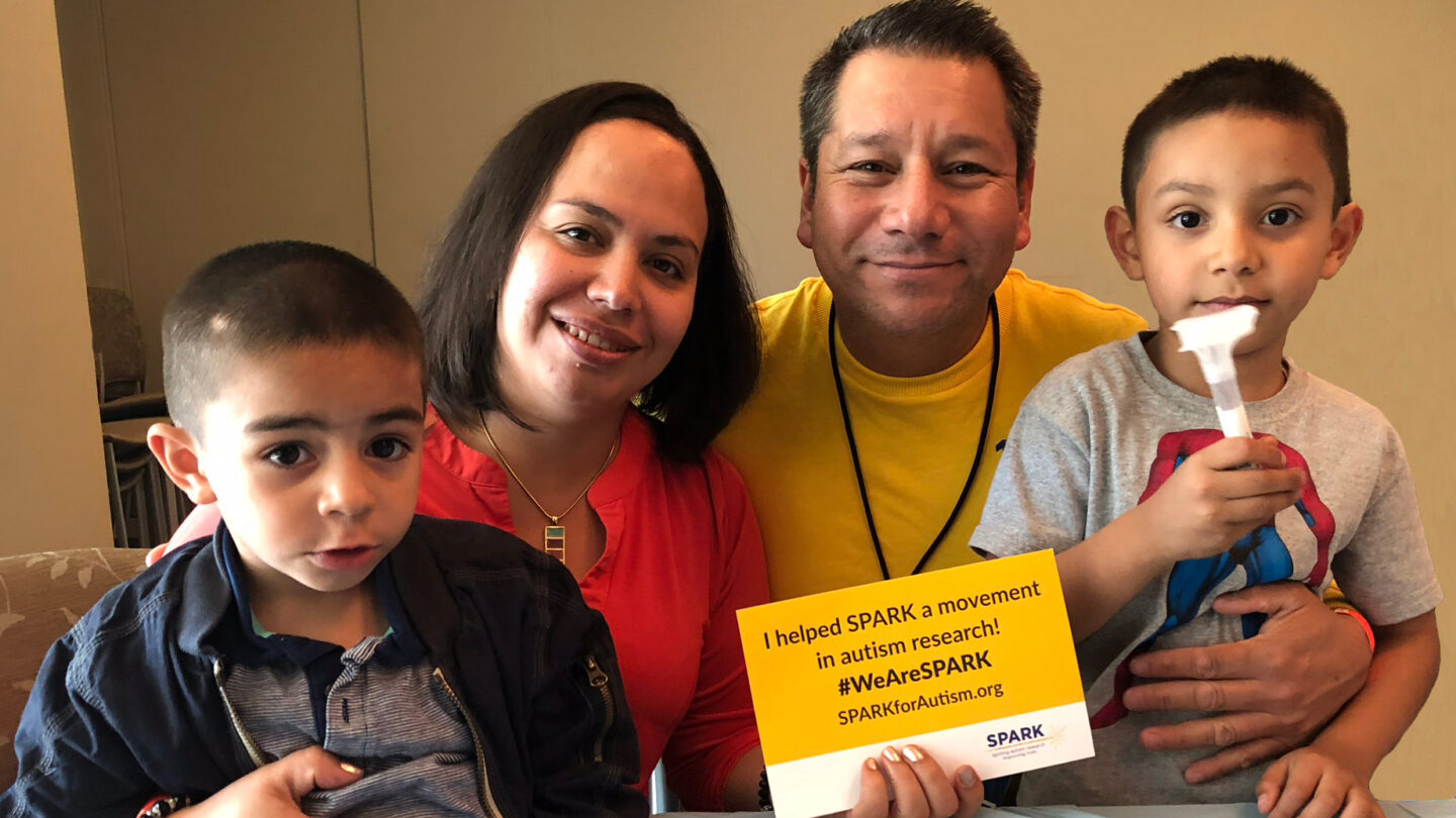 A family of four sits together smiling. An adult holds a yellow sign supporting autism research. One child holds a white object. Everyone looks happy and supportive.