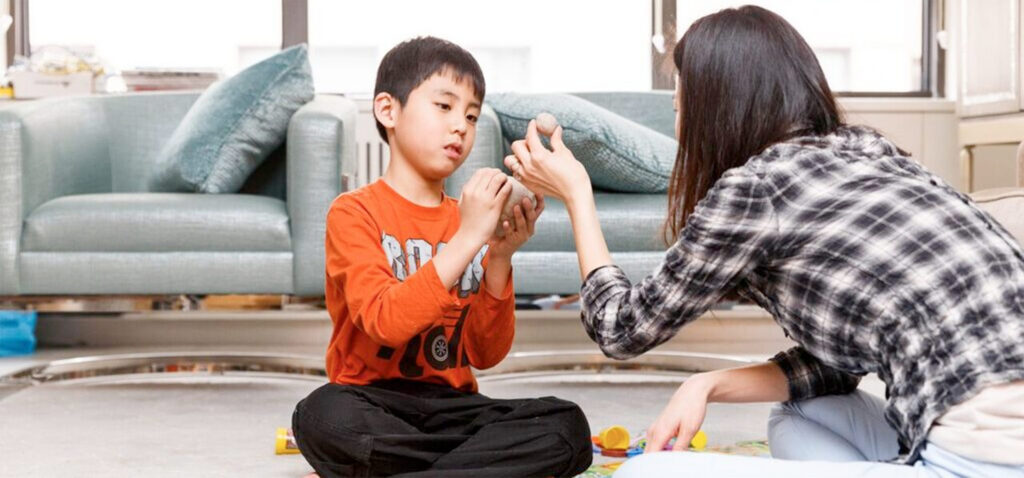 A boy in an orange shirt and an adult sit on the floor, playing with clay or dough together in a bright living room.