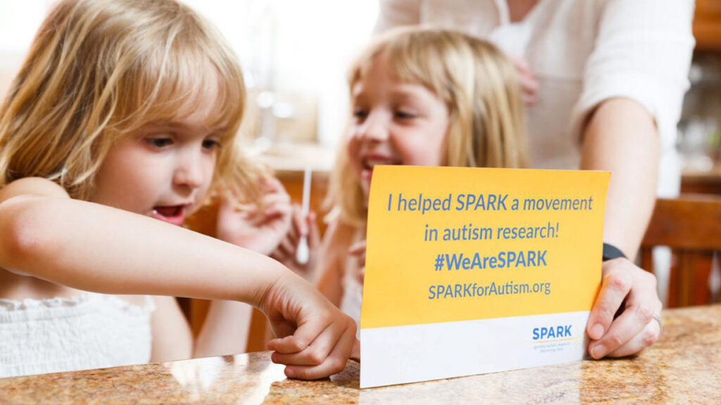 Two young children smiling and interacting with a sign that reads, I helped SPARK a movement in autism research! #WeAreSPARK SPARKforAutism.org while an adults hand holds the sign.