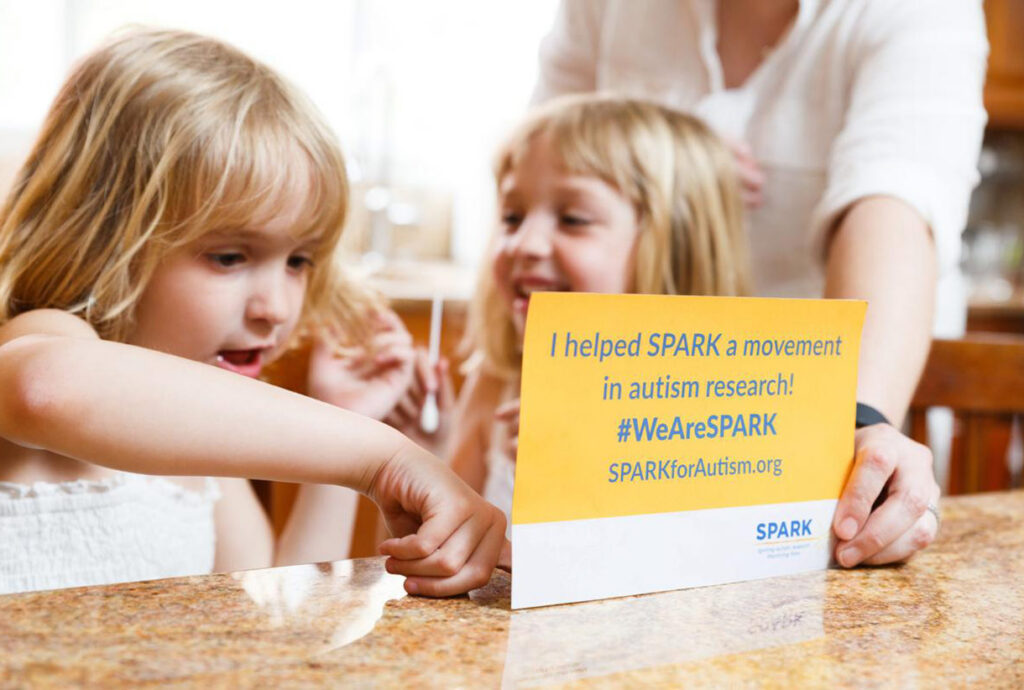 Two young children sit at a table, smiling and holding a sign that reads, I helped SPARK a movement in autism research! #WeAreSPARK SPARKforAutism.org. An adults hand is also visible.