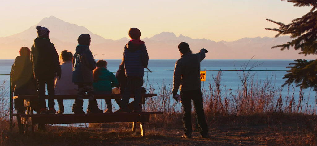 A group of people standing on and around a picnic bench overlooking the ocean and mountain range at sunset as one person points toward the horizon.