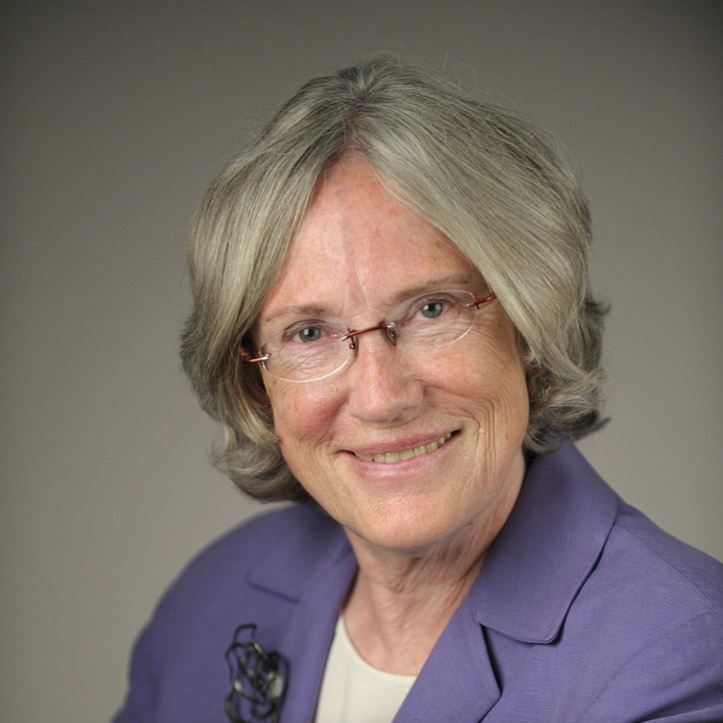 An older woman with short gray hair and glasses smiles at the camera. She is wearing a purple blazer over a light top with a brooch pinned to her lapel. The background is neutral and softly lit.